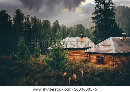 A beautiful rainy rural scenery in the mountains with shallow depth of field and selective focus on two small wooden cabins in deep taiga forest with particles of rain everywhere, dramatic stormy sky