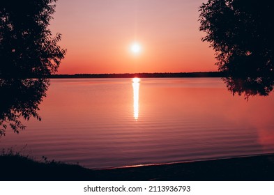 Beautiful, red bright colorful sunset on lake in frame of trees, summer landscape. Reflections, glare on rippled water.