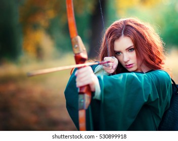 Beautiful red haired girl in green  medieval dress shooting with arrows . Fairy tale story about brave heart woman .Glowing sun on archer. Warm art work.