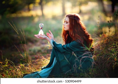 Beautiful Red Haired Girl In Green Medieval Dress In Glowing Sun Holding A Sand Glass And Looking On It. Fairy Tale Story About Brave Heart Woman.Warm Art Work