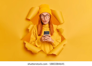 Beautiful red haired millennial woman in stylish yellow hat and jumper uses modern cellphone for online communication looks aside with wonder poses through paper hole types message to boyfrined