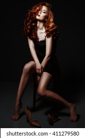Beautiful red haired woman relaxing in black silk peignoir, take off her shoes over black background. 