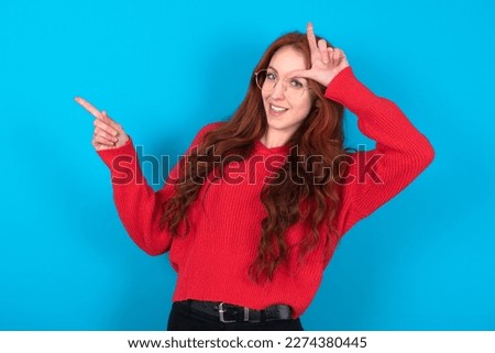 beautiful red haired woman wearing red knitted sweater over blue background showing loser sign and pointing at empty space