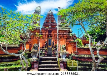 Beautiful red hindu temple gate, green trees, blue sky, mythological figures - Pura Batuan, Ubud, Bali 