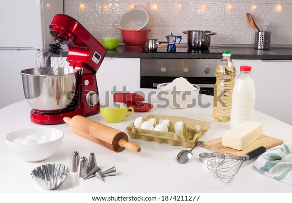 A beautiful red\
stand mixer with a metal cup stands in the kitchen. Utensils for\
backing or cooking on a table. Eggs, milk, butter, rolling pin,\
whisk, flour, vegetable oil.