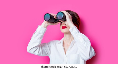 Beautiful red-haired businesswoman with binoculars on pink background