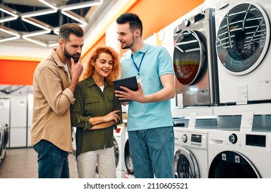 A beautiful red-haired girl and a bearded man buy a washing machine in a home appliances and electronics store. A male salesman helps a couple choose new appliances for their home. - Shutterstock ID 2110605521
