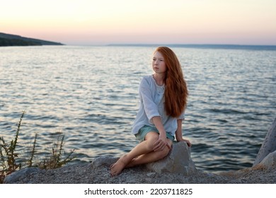 A Beautiful Red-haired Girl Sits On A Stone On The Banks Of A River, Lake, Sea. She Is Looking At Ocean And Thinking Dreamily. Girl Alone Outside. Girl Sitting On Rocks. Lonely Person. 