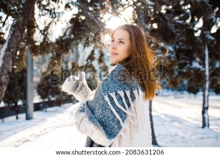 A beautiful red-haired girl in a woolen sweater walks in a snowy park in winter. A close-up shot of a woman on a sunny winter day