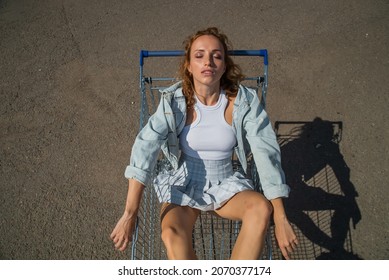 Beautiful Red-haired Model Girl In Jeans And A Short Skirt In The Parking Lot Inside Lies In A Metal Trolley For Groceries, Supermarkets, . Sexy Look Of A Woman Waiting For Top Of View-