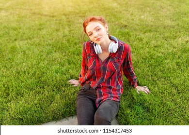 A beautiful red-haired student with freckles is dressed in a red checkered shirt with headphones sitting on the lawn in between the cheba. Student leisure.