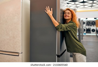 Beautiful red-haired woman hugging a refrigerator in a home appliances and electronics store. Buying a new refrigerator.
