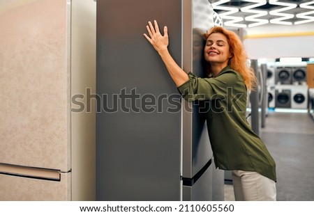 Beautiful red-haired woman hugging a refrigerator in a home appliances and electronics store. Buying a new refrigerator.