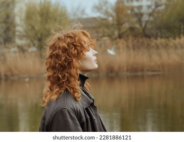 a beautiful red-haired woman walks in the park near the lake in a leather jacket, enjoying a pleasant day - Powered by Shutterstock