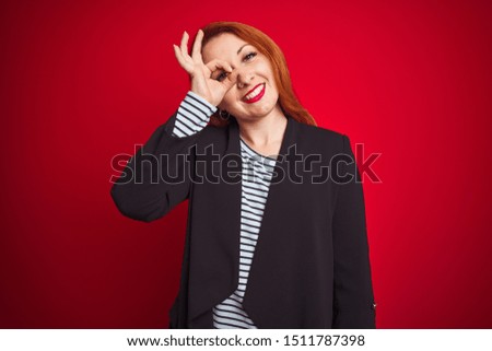 Beautiful redhead business woman wearing elegant jacket over isolated red background doing ok gesture with hand smiling, eye looking through fingers with happy face.