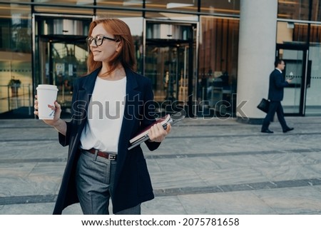 Beautiful redhead female entrepreneur standing outdoors with coffee cup and laptop in hands, leaving office, businesswoman in spectacles dressed in formal clothes waiting for taxi and looking aside