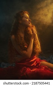 Beautiful Redhead Freckled Woman With Long Natural Curly Hair Wearing Red Dress Posing In Smoke, Darkness And Warm Light, Smoke, Sunny Rays. Art Portrait
