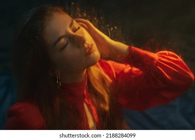 Beautiful Redhead Freckled Woman Wearing Red Dress Posing In Smoke, Darkness And Warm Light. Art Portrait
 Through The Wet Glass
