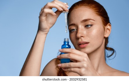 Beautiful Redhead Girl Applying Vitamin Serum On Face. Young Woman With Red Hair Open Blue Bottle Of Skin Care Product, Holding Dropper With Cosmetic Serum, Isolated On Studio Background