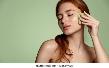 Beautiful redhead woman doing skin care routine after shower, scrapping, massage facial skin with jade gua sha scrapper, body and face beauty concept, green background.