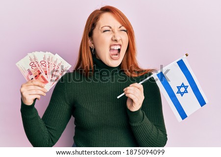Beautiful redhead woman holding 20 shekels banknotes and israel flag angry and mad screaming frustrated and furious, shouting with anger. rage and aggressive concept. 