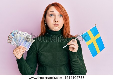 Beautiful redhead woman holding 20 swedish krona banknotes and sweden flag puffing cheeks with funny face. mouth inflated with air, catching air. 