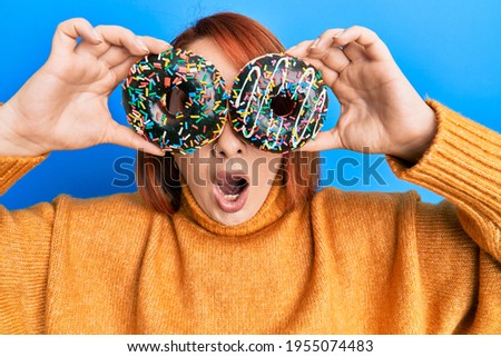 Beautiful redhead woman holding tasty colorful doughnuts on eyes afraid and shocked with surprise and amazed expression, fear and excited face. 