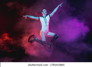 Beautiful redhead woman in sportswear jumping in a kangoo jumps shoes isolated on dark gradient studio background in neon lighted smoke. Active movement, action, fitness and wellness. Slender model.