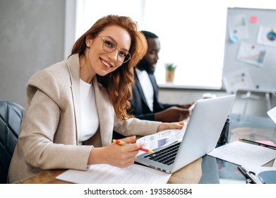 Beautiful redhead woman is using laptop, working on project with colleagues. Happy female employee in stylish suit is sitting in modern office on business meeting, looking at the camera, smiling - Powered by Shutterstock