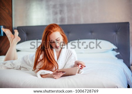 beautiful redhead woman wearing bathrobe and using digital tablet while lying on bed