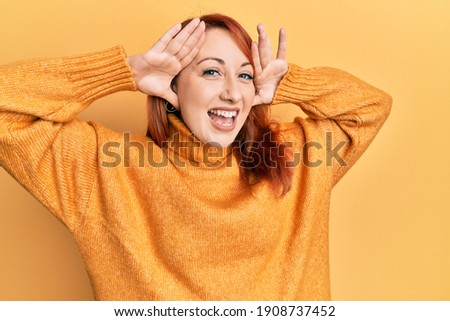 Beautiful redhead woman wearing casual winter sweater over yellow background smiling cheerful playing peek a boo with hands showing face. surprised and exited 
