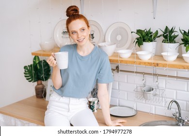 Beautiful Redhead Young Woman Holding Cup Of Hot Coffee While Standing In Kitchen Room With White Modern Interior. Smiling Business Lady Drinking Tasty Beverage From Mug At Home Office In During Break