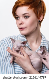 Beautiful Redhead Young Woman Hugging Sphynx Cat And Looking Away. Studio Shot On White Background. Portrait Of Hipster With Short Hair Dressed In Striped White-blue Casual Shirt. Part Of Series.