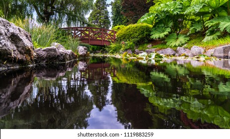 Beautiful Reflexion Of A Wooden Bridge Above  Water Can Flowers And Rocks On A Smooth Water Surface.