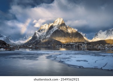 Beautiful reinebringen mountain towers over fishing village Reine in Lofoten islands, Norway, with partially frozen fjord and rainbow in winter at sunset. Dramatic sky over snowy rocks and sea. Travel - Powered by Shutterstock