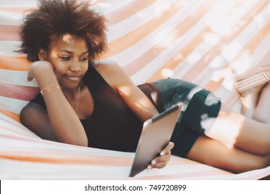 Beautiful Relaxed Black Female Student With Curly African Hair Is Laying On Striped Hammock In Campus And Reading Her Homework Tasks On The Screen Of Digital Tablet On Sunny Summer Morning