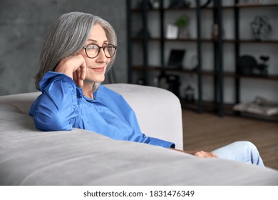 Beautiful Relaxed Mature Old Woman Relaxing Sitting On Couch At Home. Peaceful Middle Aged Grey-haired Lady Wearing Glasses Resting On Sofa Enjoying Lounge, No Stress, Peace Of Mind, Looking Away.