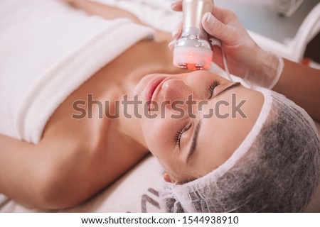 Beautiful relaxed woman enjoying rf-lifting session at beauty salon. Professional cosmetologist doing skin tightening procedure for female client