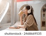 A beautiful, relaxed young Asian woman is gazing at the view outdoors and daydreaming while sipping coffee and listening to music on her headphones in a cafe. leisure, enjoyment, happiness