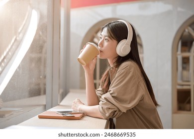 A beautiful, relaxed young Asian woman is gazing at the view outdoors and daydreaming while sipping coffee and listening to music on her headphones in a cafe. leisure, enjoyment, happiness - Powered by Shutterstock