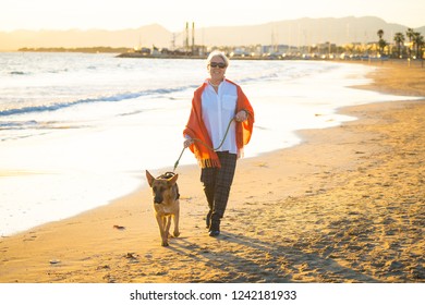 Beautiful Retired Older Woman And Pet German Shepard Dog Walking Along The Shore Sea Ocean On Beach In Companionship Benefits Of Animals Keeping Active Retirement Lifestyle And Dog Friendly Tourism.