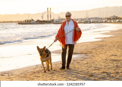 Beautiful Retired Older Woman And Pet German Shepard Dog Walking Along The Shore Sea Ocean On Beach In Companionship Benefits Of Animals Keeping Active Retirement Lifestyle And Dog Friendly Tourism.