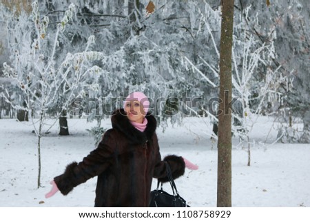 Beautiful and rich middle-aged woman in fur coat and pink winter hat is joking, smiling, and having fun in the snow-covered park.