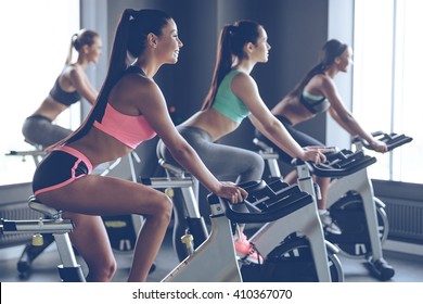 Beautiful ride. Side view of young beautiful women with perfect bodies in sportswear looking away with smile while cycling at gym