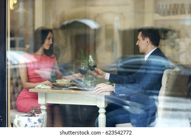 Beautiful Romantic Couple Having Dinner With Wine In Bright Luxury Restaurant. View Through Window From Outside