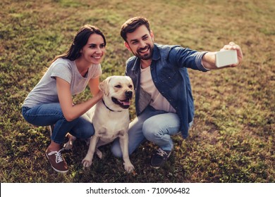 Beautiful Romantic Couple Is Having Fun With Their Dog Labrador Retriever Outdoors. Sitting On A Green Grass And Making Selfie On A Smart Phone.