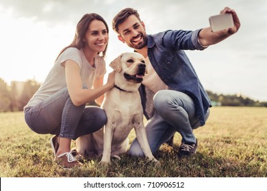 Beautiful romantic couple is having fun with their dog labrador retriever outdoors. Sitting on a green grass and making selfie on a smart phone.