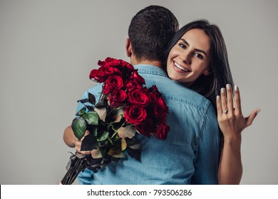 Beautiful Romantic Couple In Love Isolated On Grey Background. Attractive Young Woman With Ring On Hand Is Holding Red Roses While Hugging Her Handsome Man. Just Engaged. Happy Saint Valentine's Day!