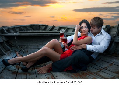 Beautiful Romantic Couple With Wineglasses Of Red Wine In Old Boat On Sunset Background