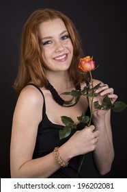 Beautiful romantic happy woman in elegant black evening wear holding a single long stemmed rose from her sweetheart and smiling at the camera on a dark background.